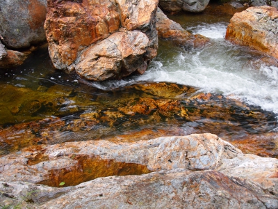 [Shallow water with a tan and black patterned rock bottom visible through it. Surrounding it are dry grey and salmon colored rock.]